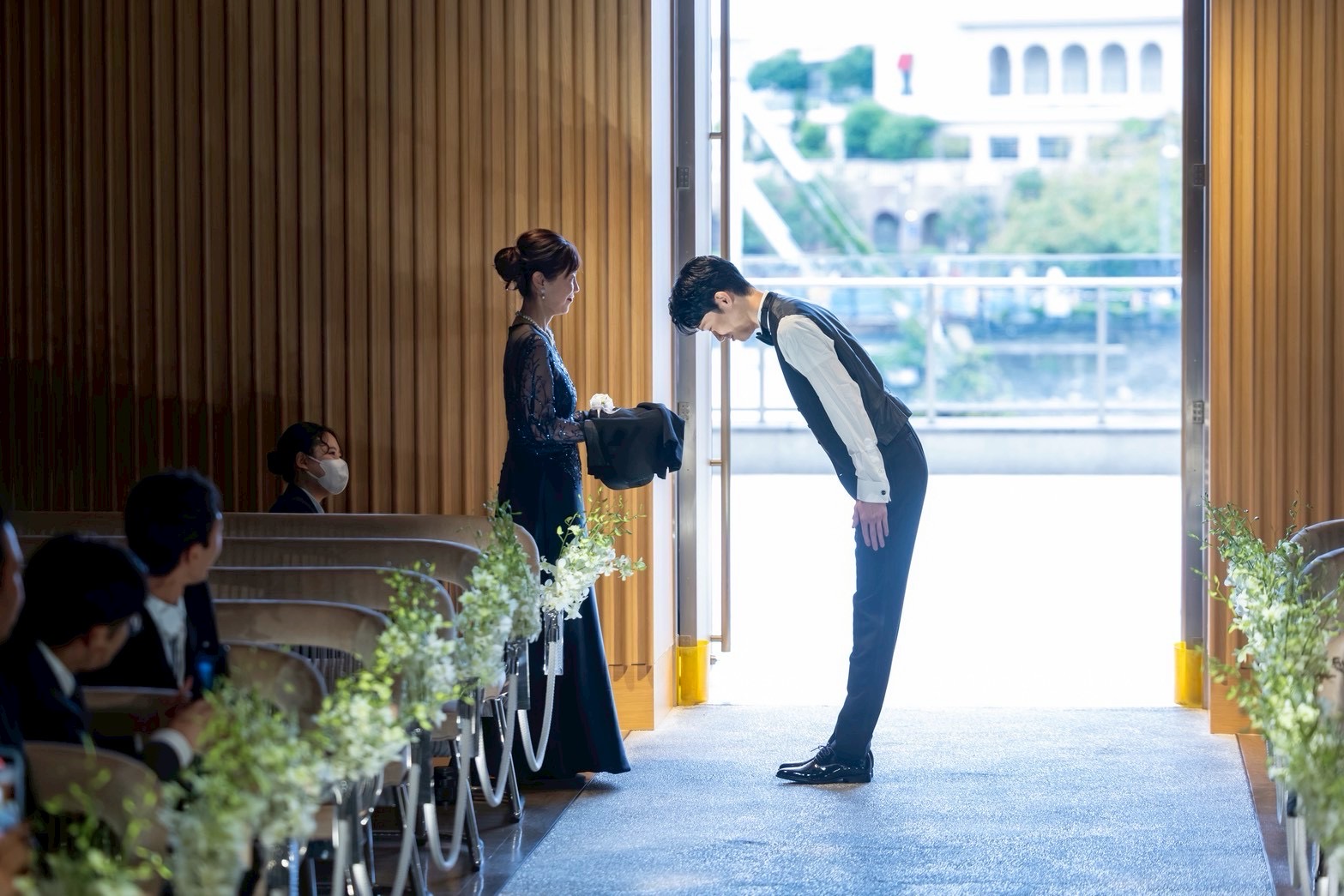 ジャケットセレモニーで新郎が母に深くお辞儀をする感動的な結婚式シーン。ネイビーのドレス姿の新郎母が見守る厳かな瞬間。