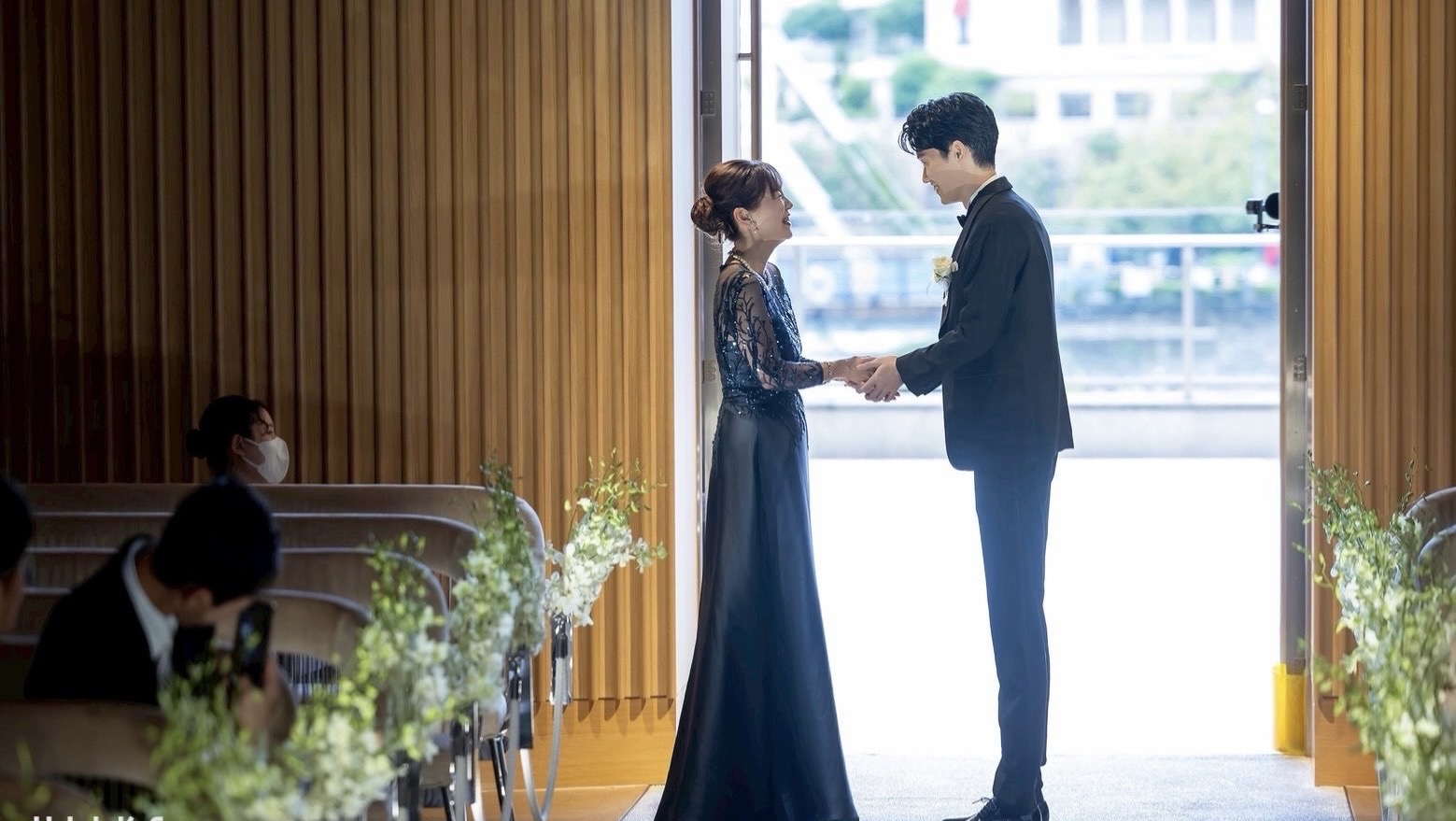 新郎母がネイビーのビジュードレスを着て、ジャケットセレモニーで新郎と手を取り合う感動的な瞬間。自然光に包まれた結婚式のワンシーン。