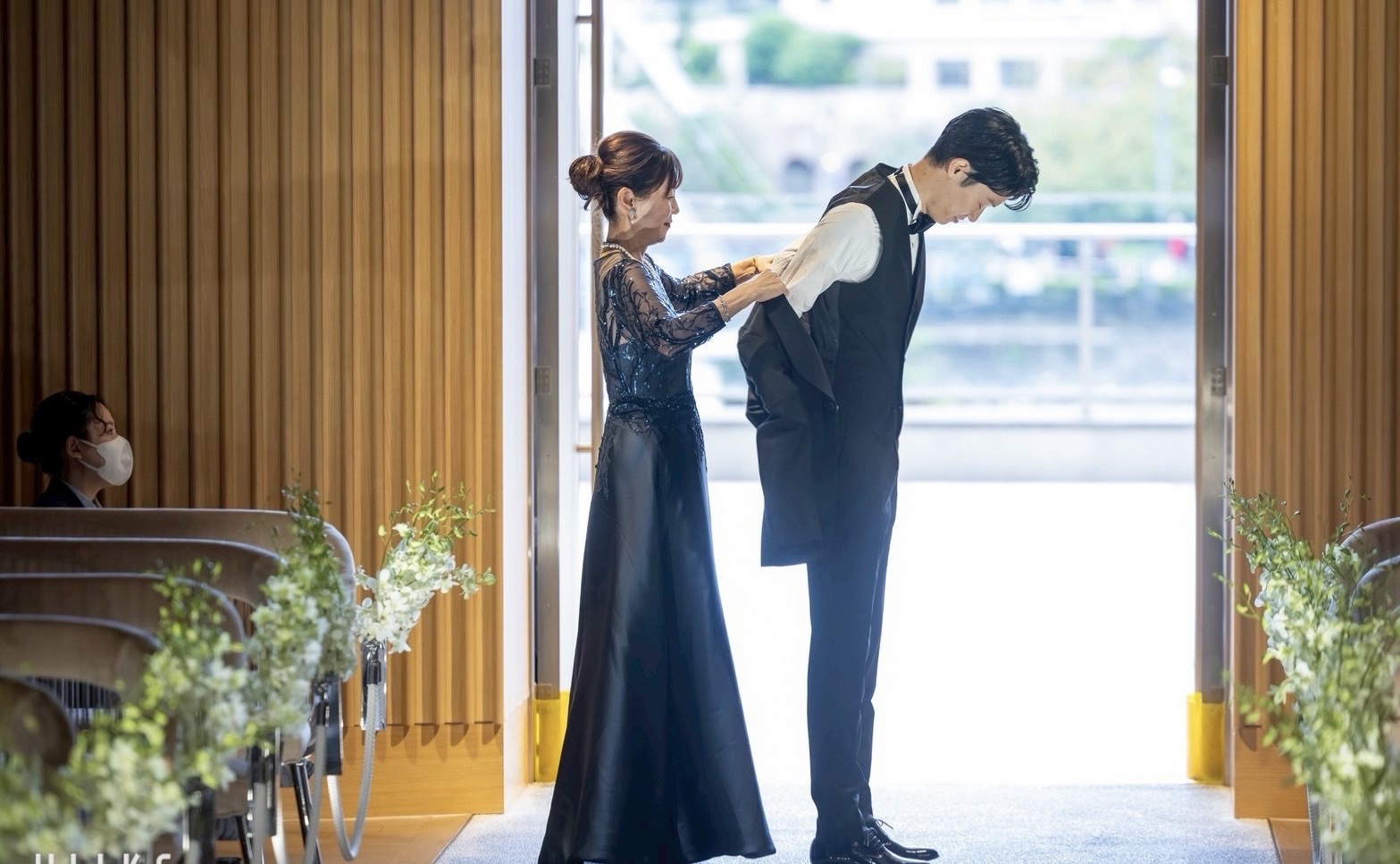 新郎母がネイビーのトレーンドレスを着用し、ジャケットセレモニーで新郎にジャケットを着せる感動的な瞬間。横から見た美しいAラインシルエット。