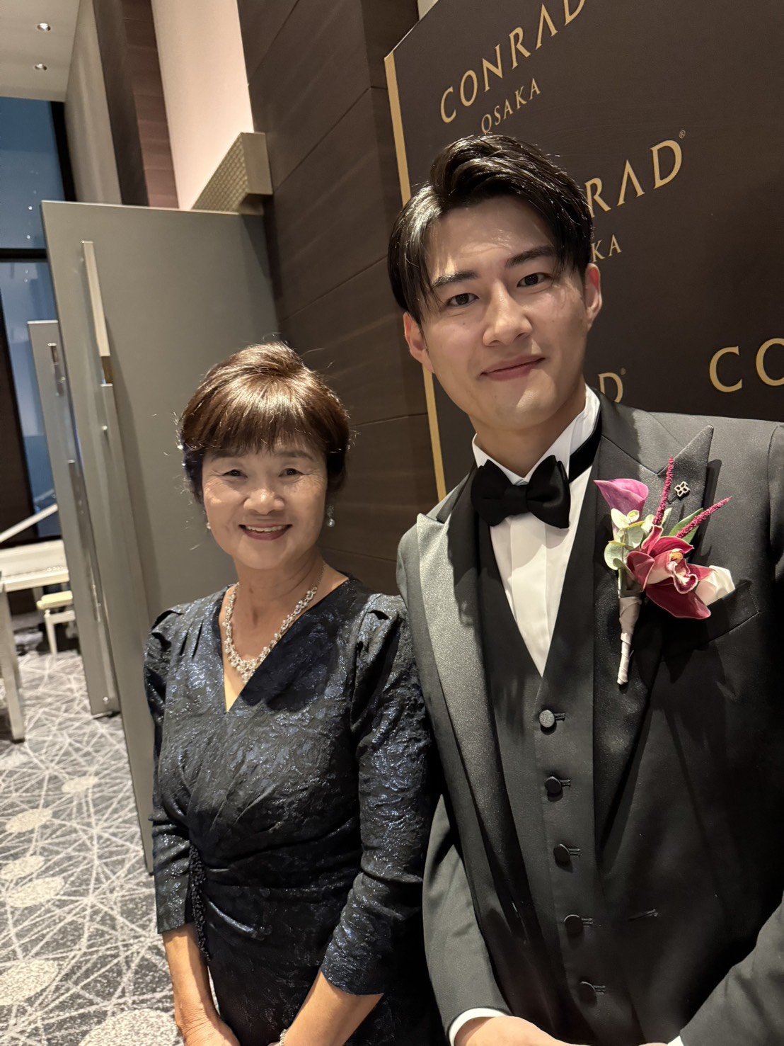 ラメ感のあるネイビードレスを着た新郎母と、ブラックタキシードの新郎が並ぶツーショット。Vネックと七分袖の上品なデザインが際立つフォーマルな装い。