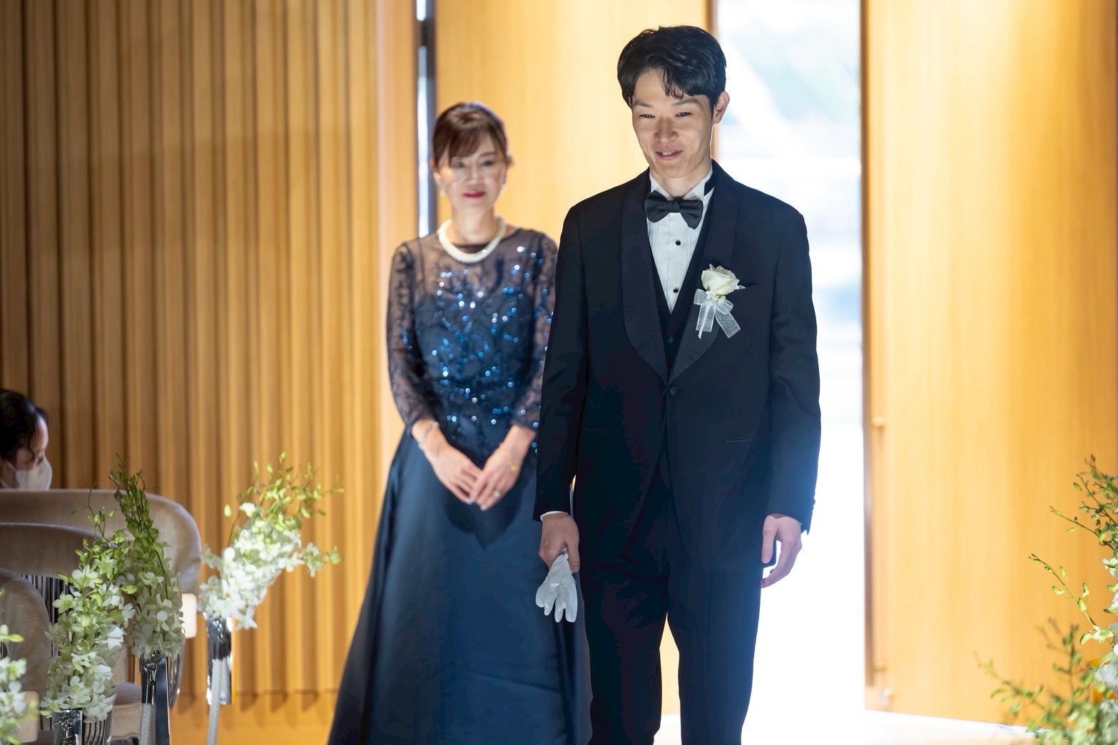 結婚式でセレモニー後に新郎が歩み出し、ネイビーのスパンコール刺繍ドレスを着た母が温かく見守る感動的なシーン。光を受けてドレスが上品に輝く。