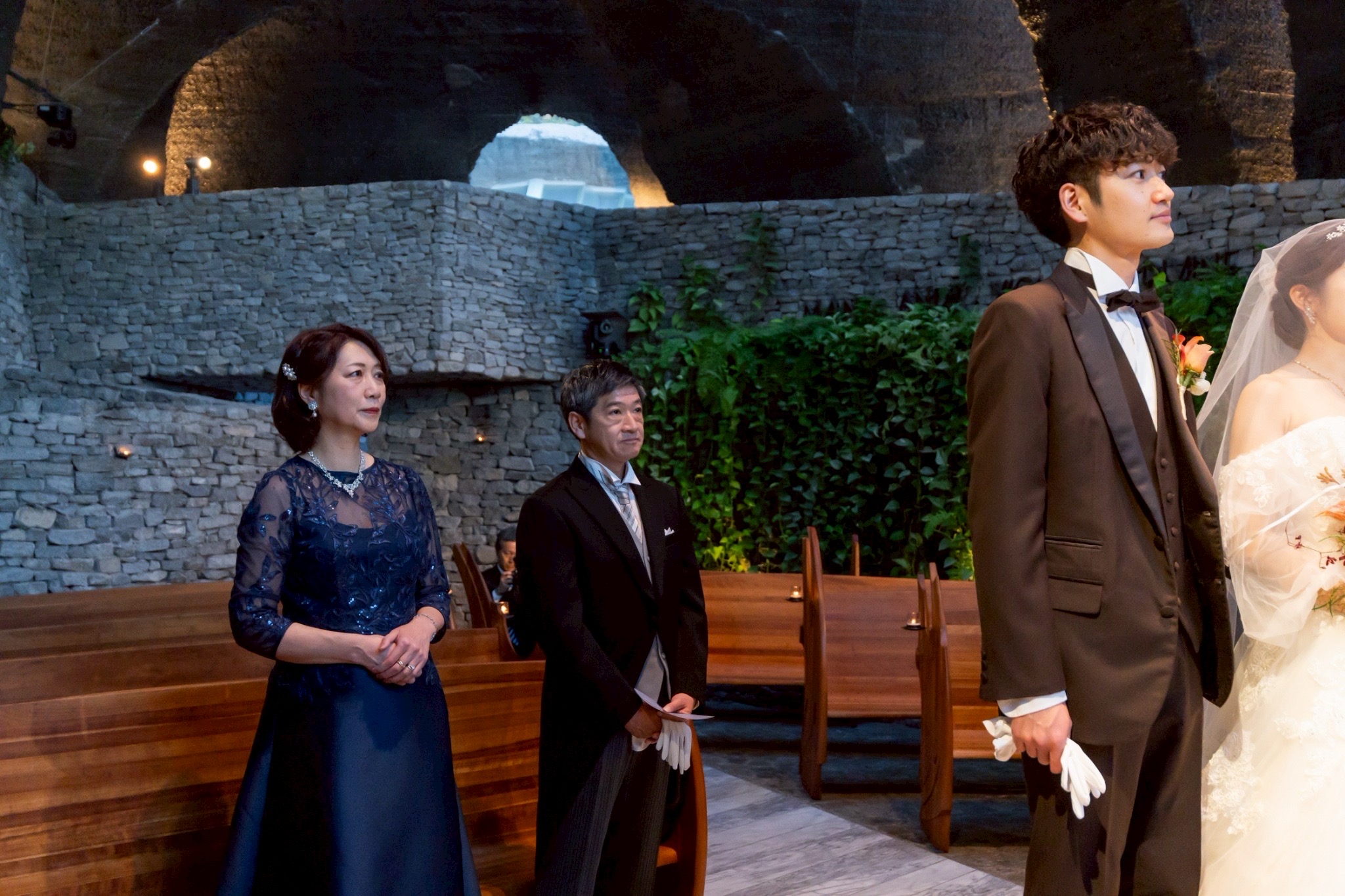 結婚式のチャペル内で、新郎と両親が並ぶフォーマルなシーン。ネイビーのマザードレスとモーニングコートの親族コーディネート。