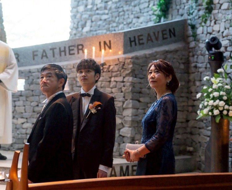 石造りのチャペルでの結婚式中、新郎と両親が並ぶシーン。新郎母はネイビーのレースドレスを着用し、上品でフォーマルな装い。