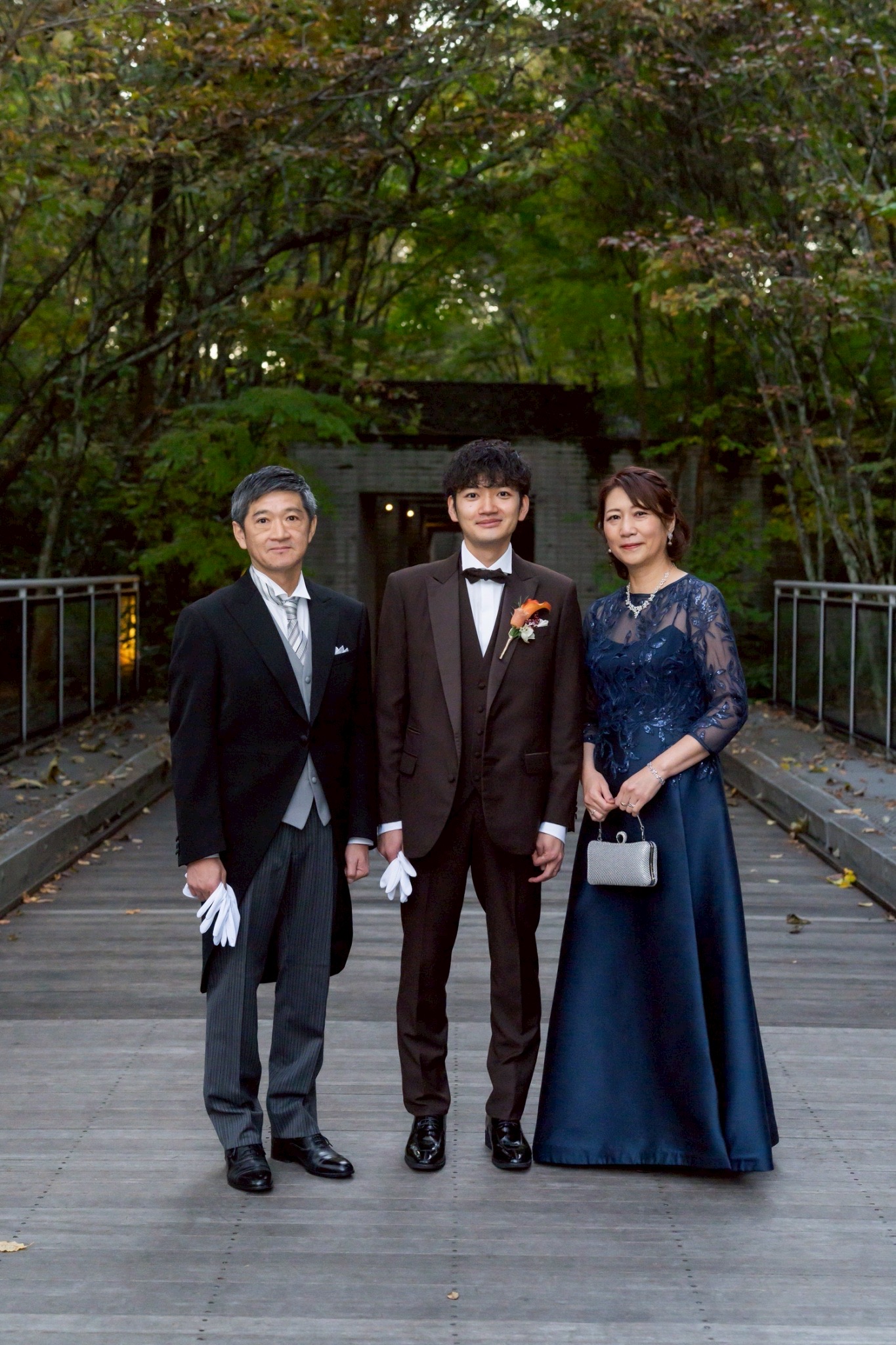 軽井沢の秋の自然の中で、新郎と両親が並ぶ結婚式の家族写真。新郎母はネイビーのレースドレスを着用し、上品で落ち着いたフォーマルコーディネート。
