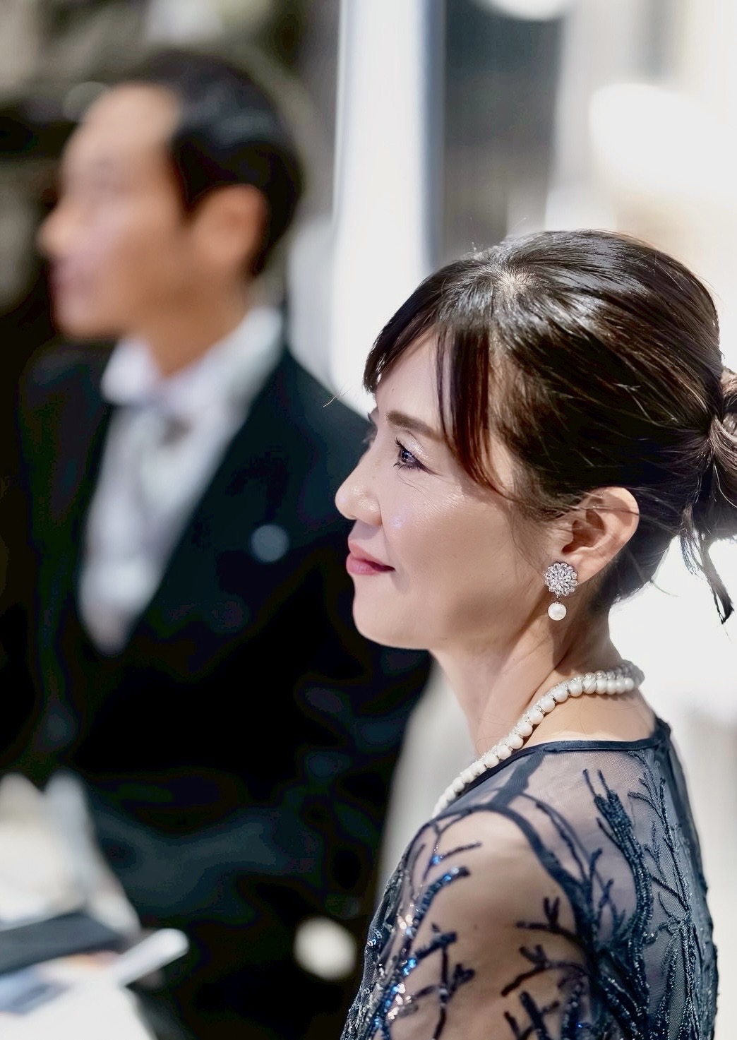 ネイビーのレースドレスを着た新郎母の横顔アップ。上品なアップヘアとパールアクセサリーを合わせ、高砂を見つめる穏やかな表情の結婚式シーン。