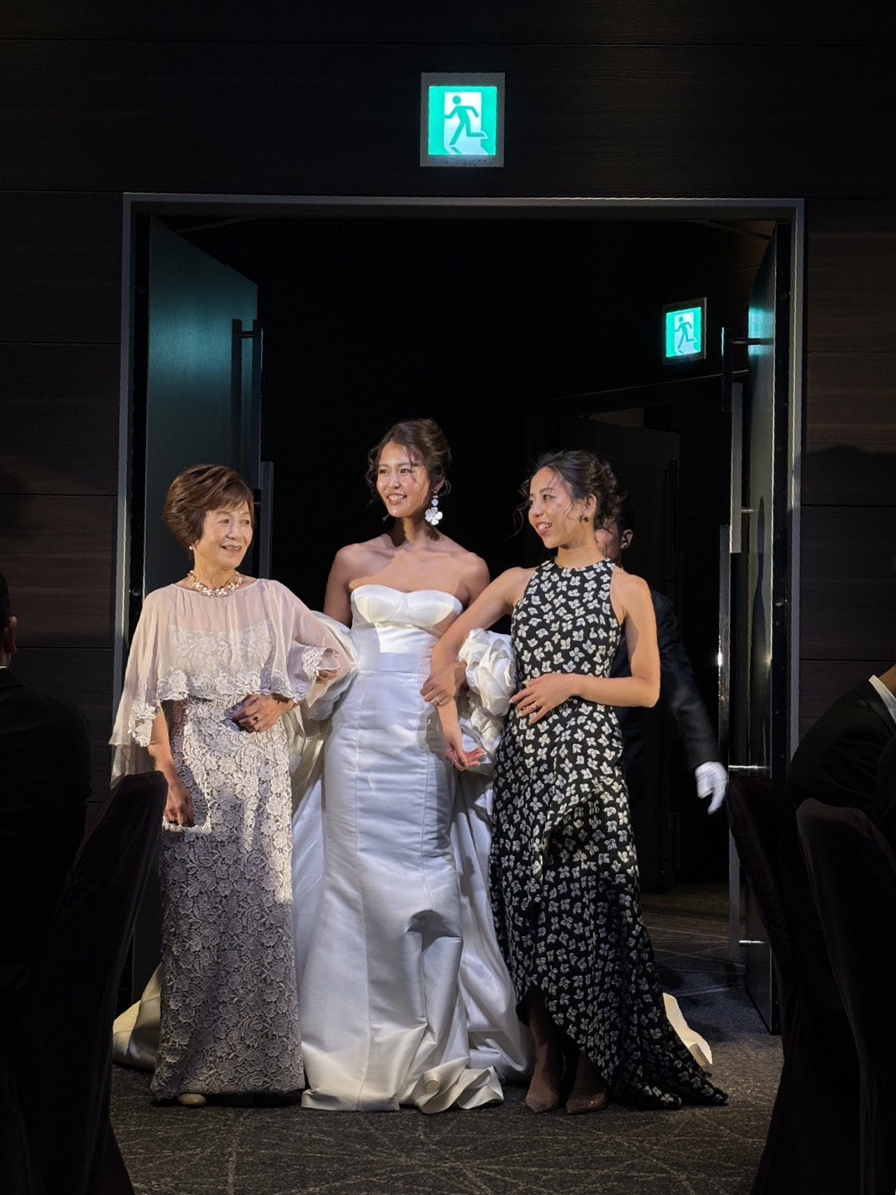披露宴の中座で新婦と新婦母、新婦姉が腕を組んで歩くシーン。レースケープのマザードレスとスタイリッシュなドレス姿の姉妹の華やかなドレスアップ