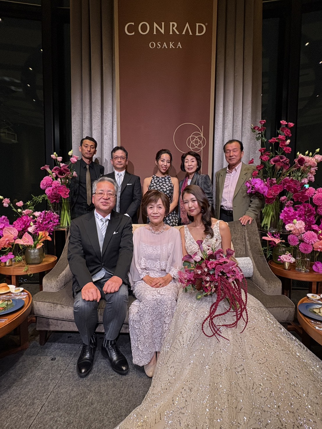 新婦母を中心に新郎新婦が並び、後ろに新婦父やご親族が立つ結婚式の家族集合写真。上品なレースケープのマザードレス姿の新婦母と笑顔あふれるご家族